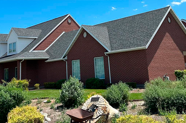 A red brick house with gray shingle roofing, two large windows, a landscaped front yard, and a wheelbarrow in the foreground.