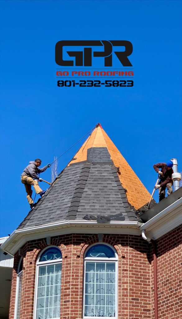 Two workers install shingles on a steep, conical roof of a brick building under a clear blue sky. The "Go Pro Roofing" logo and contact number are displayed above.