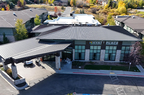 Aerial view of a Hyatt Place hotel entrance with covered driveway, large windows, stone accents, and surrounding trees.