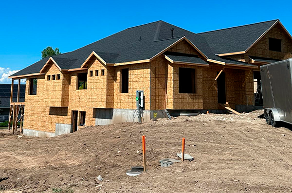 A house under construction with plywood walls and a black shingled roof, surrounded by dirt and construction equipment.
