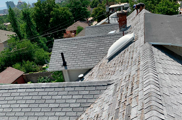 A person is repairing shingles on a sloped residential roof, with other rooftops, trees, and city buildings visible in the background.