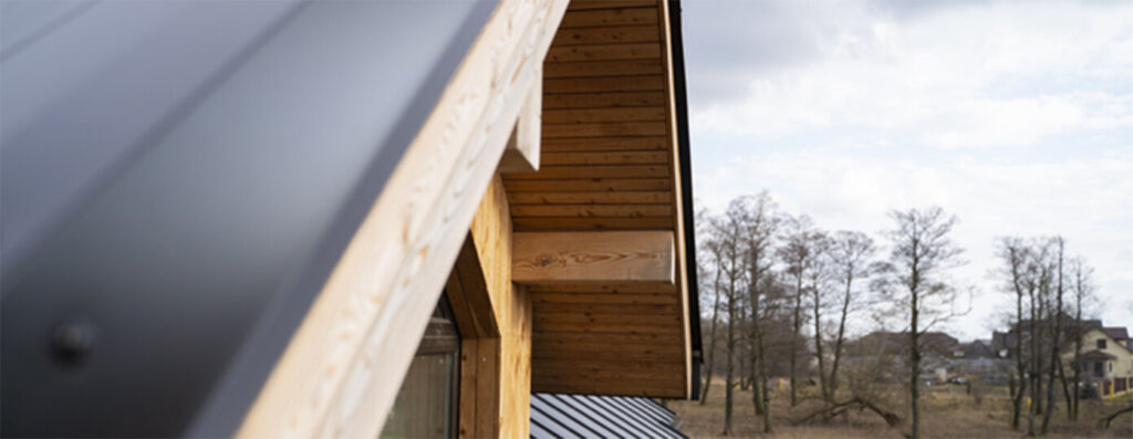 Close-up view of a modern wooden house roof with metal sheeting, with bare trees and houses visible in the background under a cloudy sky.